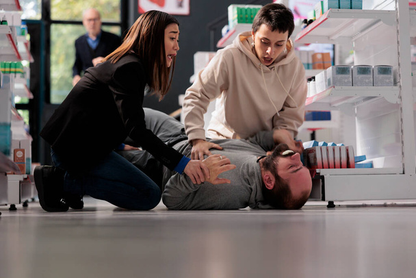Women standing on knees near unconscious man with epilepsy lying on pharmacy retail store floor. Young person suffering from foaming ar mouth and convulsions neurological disorder symptoms - Foto, afbeelding