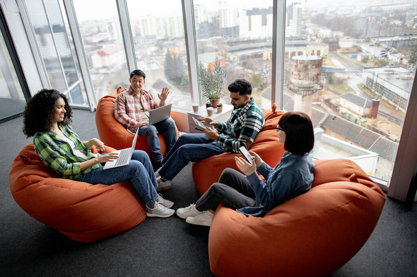 Diverse group of professionals using smart gadgets while resting in circle of soft seats in creative workspace. Multiracial managers in street style clothes planning e-commerce investment strategy. - Foto, afbeelding