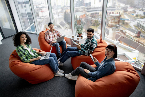 Top view of diverse male and female adults in casual clothes posing on terracotta soft seats with digital gadgets. Multiethnic collaborators planning presentation for colleagues in shared workspace. - Fotografie, Obrázek