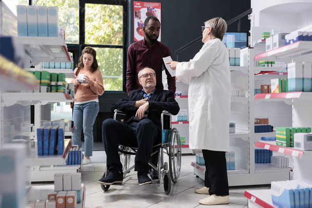 Huoltaja auttaa vanhempi asiakas pyörätuolissa terveydenhuollon ostoksia apteekissa, joka tarjoaa reseptiä apteekkiin pyytää apua pillereitä. Lääketieteellinen palvelu ja tuki - Valokuva, kuva