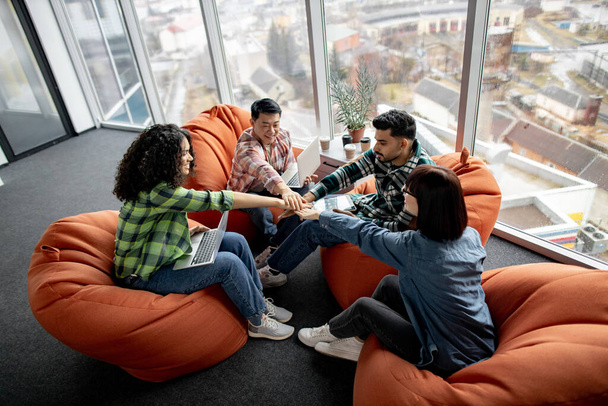 Four multicultural businesspeople in street style outfits stacking hands while gathering in tight circle in coworking space. Smiling colleagues with devices participating in team building activity. - Foto, Imagen