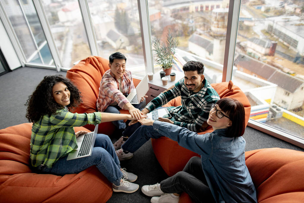 Group of happy young architects posing on beanbags while huddling in circle with hands together in creative office. Successful multiethnic partners with gadgets succeeding common business goal. - 写真・画像