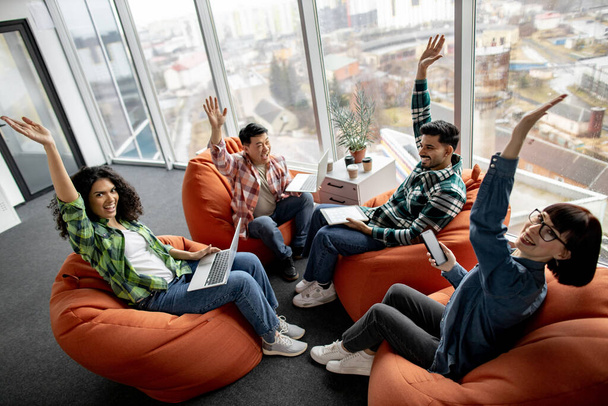 Excited male and female employees putting hands in air after stacking them while working with mobile devices in meeting space. Multiethnic designers in casual outfits motivating team spirit indoors. - Foto, afbeelding