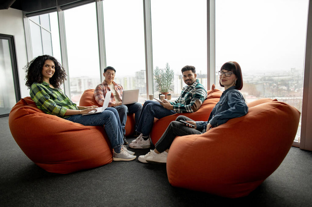 International men and women in everyday wear posing with handheld gadgets while resting on pouf chairs in creative workplace. Successful colleagues boosting business efficiency via teamwork benefits. - Zdjęcie, obraz
