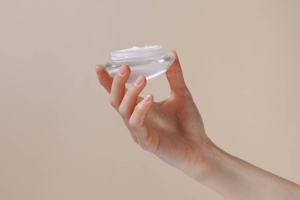 Mão feminina segurando um frasco de creme hidratante branco em um fundo isolado bege. Cuidado com a pele sensível com extrato natural. Conceito de cuidados corporais de verão ou outono - Foto, Imagem