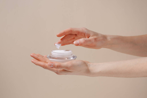 Female hand applying white moisturizing cream from glass jar on finger on beige isolated background. Concept of beauty, cosmetic product, skin treatment and protection - Photo, Image