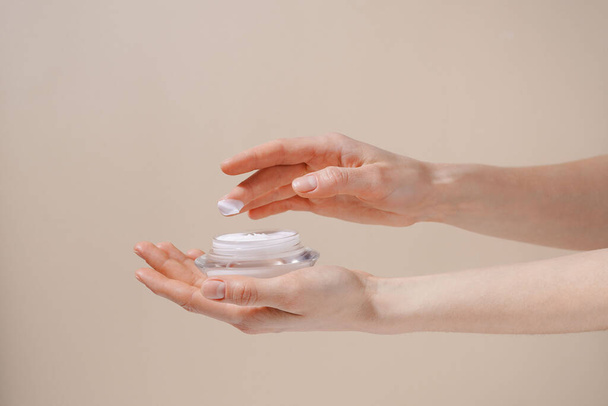 Weibliche Hand, die weiße Feuchtigkeitscreme aus einem Glasgefäß auf dem Finger auf beigem Hintergrund aufträgt. Schönheits-, Hautpflege- und Ästhetikkonzept - Foto, Bild