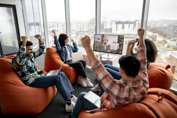 Excited businesspeople cheering chief executive of online briefing via interactive display in professional space. Multiracial colleagues celebrating company achievements on internatinal trade market. - Фото, зображення