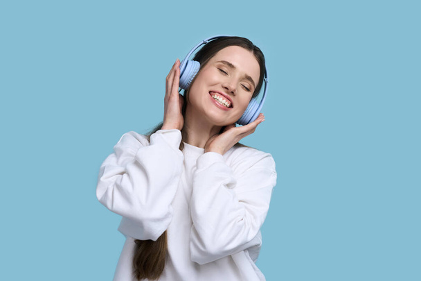 Charming stylish young Caucasian woman with wireless headphones, enjoying listening to a new cool soundtrack and dancing to music, isolated over blue background. People. Entertainment. Hobby. Leisures - Zdjęcie, obraz