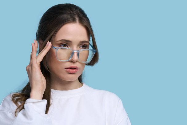 Kavkazská mladá žena ve stylových brýlích s modrým rámečkem, hledící na prázdné místo pro reklamní text, izolovaná na modrém pozadí. Moderní brýle. Zrak. Vize. Móda. Oftalmologie - Fotografie, Obrázek