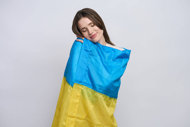 Piękna ukraińska młoda patriotka zawinięta w ukraińską flagę, delikatnie ją przytulająca, uśmiechnięta, pozująca z zamkniętymi oczami nad odizolowanym białym tłem. Koncepcja wspierania Ukrainy i powstrzymania wojny - Zdjęcie, obraz