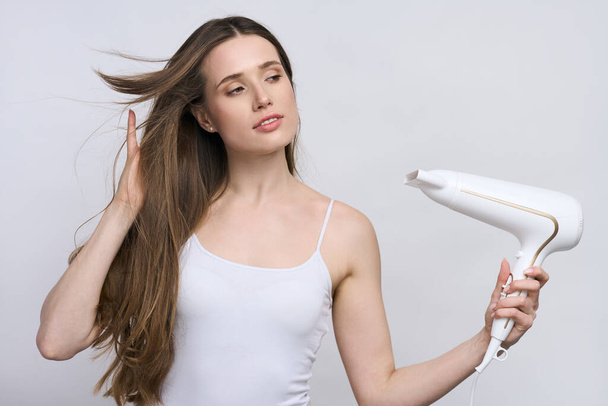 Isolated portrait on white background of Caucasian confident young pretty woman, wearing white top, holding stylish modern electric hair dryer and blowing warm air on her beautiful silky long wet hair - Photo, Image