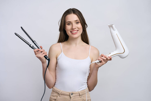Caucasian young beautiful woman holding hair styling instruments, a hair straightener and dryer, smiling looking at camera isolated on white background. Hairdresser. Barber. Style and fashion concept - Foto, afbeelding