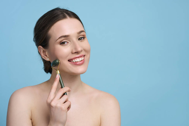 Attractive Caucasian young woman smiles looking at camera, performs lymphatic drainage or face lifting massage with jade stone roller massager, posing with naked shoulders on blue isolated background - Фото, изображение