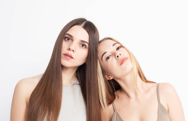 Two young women with long hair stand together on a white background, pose, look at the camera, lifestyle concept. - Photo, image