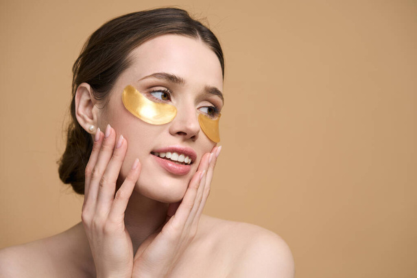 Caucasian young pretty woman applying golden collagen patches under her eye, smiling looking aside, posing bare shoulders, isolated on beige background. Facial treatment. Cosmetology. Beauty and spa - Foto, imagen
