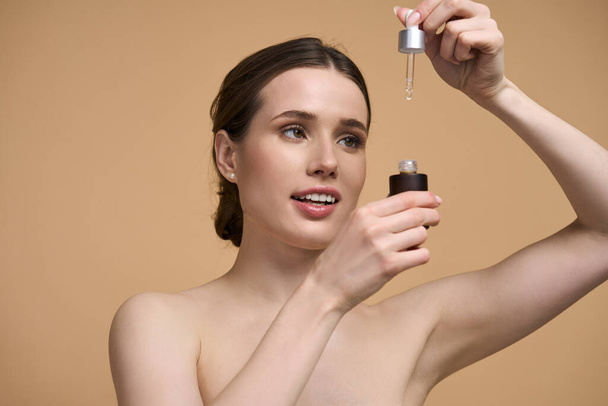 Closeup of good-looking young pretty woman, posing bare shoulders on isolated beige background, holding a bottle with anti aging serum and dripping few drops. Skin care for preventing first wrinkles - Φωτογραφία, εικόνα