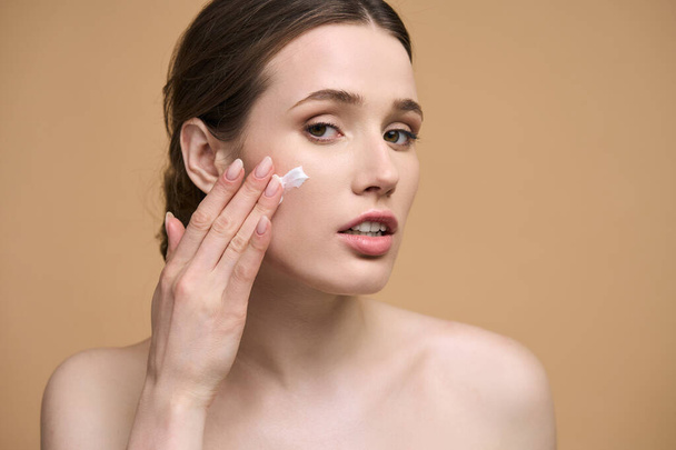 Close-up Caucasian young woman with healthy glowing clear skin, applying moisturizer on her face, looking at camera, standing with naked shoulders isolated over beige background. Beauty treatment - Photo, Image