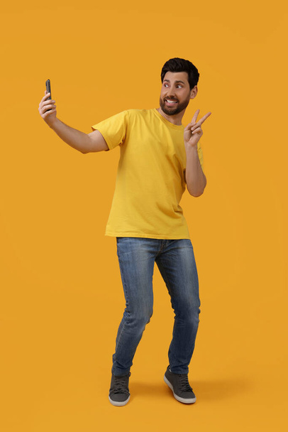 Smiling man taking selfie with smartphone and showing peace sign on yellow background - Foto, Imagen