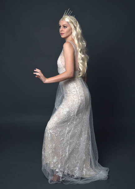 Full length πορτρέτο των όμορφων γυναικών με μακριά ξανθά μαλλιά, φορώντας στέμμα πριγκίπισσα φαντασία και κομψό λευκό φόρεμα μπάλα, στέκεται ποζάρουν με χειρονομία χέρι. Απομονωμένο σε σκούρο γκρι φόντο στούντιο. - Φωτογραφία, εικόνα