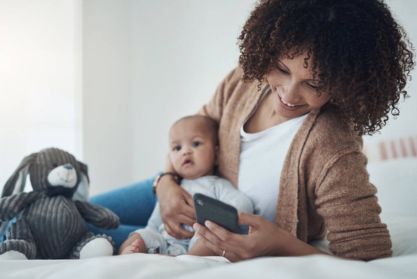 Social media is filled with moms just like you. a young woman using a smartphone while relaxing on the bed with her adorable baby girl at home - Foto, Imagem