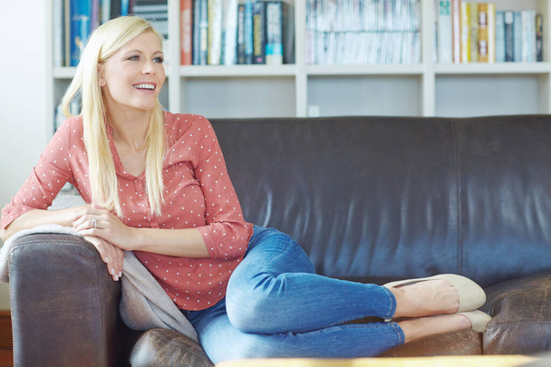 Beautiful young blonde woman daydreaming and relaxing at home on the sofa. Fullbody of one cheerful female sitting on the couch in the living room. Comfy woman smiling while resting alone. - Zdjęcie, obraz