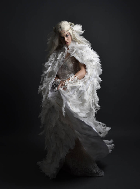 retrato de fantasía de hermosa modelo femenina con el pelo largo y rubio con traje de capa de plumas blanco de otro mundo y tocado, aislado en el fondo oscuro del estudio. - Foto, imagen
