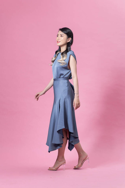 Mooie stijlvolle meisje met lange krullend haar dragen mooie blauwe jurk en poseren tegen roze achtergrond - Foto, afbeelding