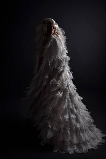 fantasy portrait of beautiful female model with long blond hair wearing otherworldly  white feathered cloak costume and headdress, isolated on dark studio background. - Photo, Image