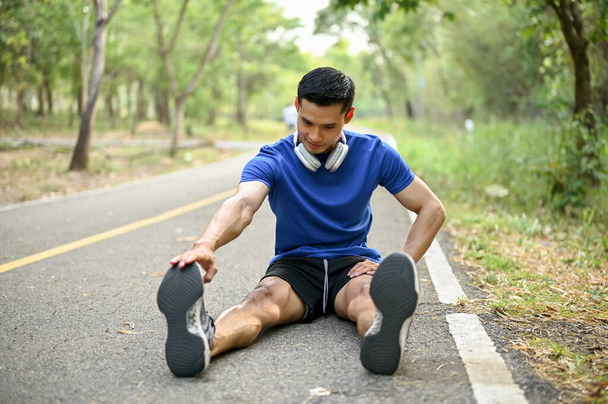 Hermoso hombre asiático milenario estirando las piernas en la calle, calentándose antes de correr en un parque natural. Estilo de vida y concepto deportivo - Foto, Imagen