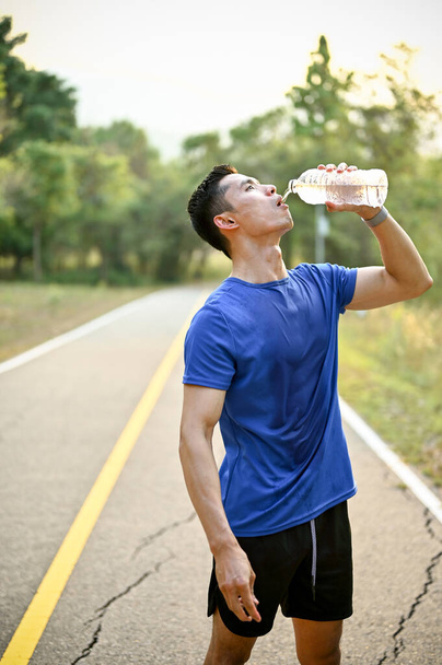 Thirsty and exhausted millennial Asian male runner drinks water from a bottle while running in a park on a sunny day. - Zdjęcie, obraz