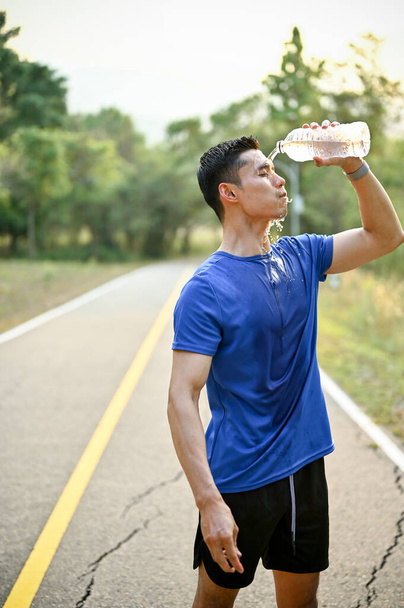 Portrait of thirsty and exhausted Asian man in sportswear pouring water from a bottle on his face after running in a park, refreshing himself by splashing water on his head. - Photo, image