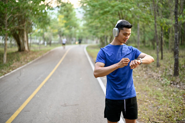 Komea tuhatvuotinen aasialainen mies käyttää kuulokkeita, tarkkailee sykettään ja juoksee kilometrejä älykellollaan juostessaan puistossa.. - Valokuva, kuva