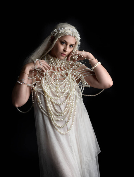 Close up portrait of beautiful blonde female model wearing an extravagant pearl jewelled gown. Gestural arms reaching out,  isolated on a black studio background. - 写真・画像