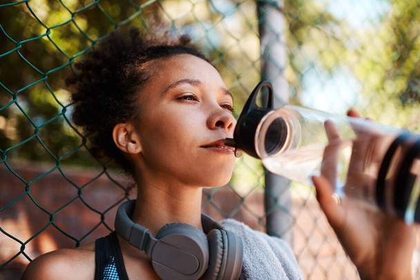 Blijven gehydrateerd helpt voorkomen dat de daling van uw prestaties. een sportieve jonge vrouw die water drinkt terwijl ze buiten tegen een hek staat - Foto, afbeelding