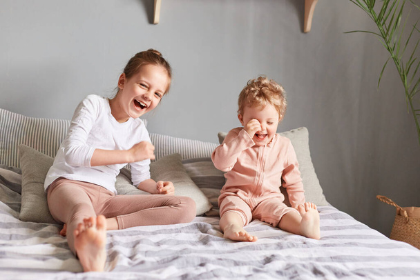 Girls or sisters at home having fun together on the bed in casual clothing laughing happily, looking at the camera, enjoying their weekend morning. - Fotografie, Obrázek