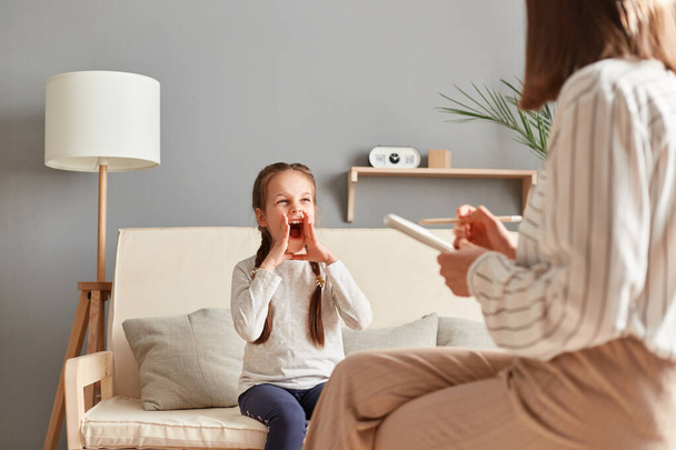 Little girl at therapy session with psychologist expressing her emotions and frustrations by shouting loudly while specialist calmly listens and takes notes. - Φωτογραφία, εικόνα