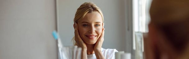 Jolie jeune femme blonde souriant dans un miroir à la maison. Salle de bains. Concept de soins de la peau. Beauté et soin. - Photo, image