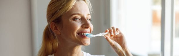 Close up. Cheerful young blonde female brushing teeth with toothbrush at mirror. Take care of teeth. - 写真・画像