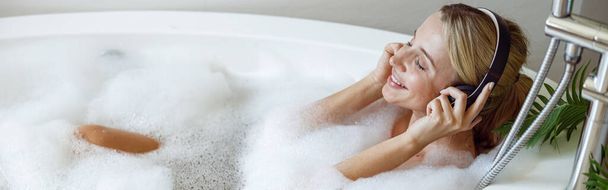 Cheerful young woman in headphones smiling, listening to music in bath with bubbles. Top view. - Foto, Imagen