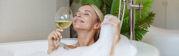 Atractiva joven feliz disfrutando de burbujas de espuma y vino blanco en la bañera de hidromasaje en el spa. Relajante. - Foto, Imagen