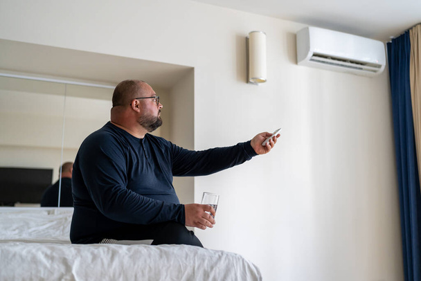 Frustrierte übergewichtige Menschen, die unter Hitze leiden, greifen zu Wasser und konditionieren sich zu Hause. Unwohlfühlende dicke Männer schwitzen, während sie auf dem Bett sitzen und versuchen, sich im Hotelzimmer abzukühlen. Heißluft in Innenräumen - Foto, Bild