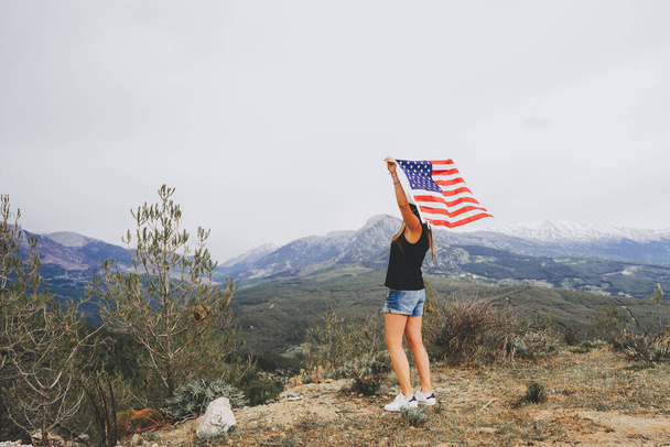 Mujer joven de pie en un acantilado de roca y ondeando la bandera de Estados Unidos mientras mira las montañas en el fondo. Chica viajera ondeando bandera americana de pie en la cima de la montaña. 4 4 de julio Día de la Independencia. - Foto, Imagen