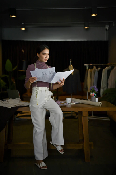 Portrait of a professional and gorgeous millennial Asian female fashion designer or tailor leaning on a table and checking her new collection sketch in her studio. - Photo, Image