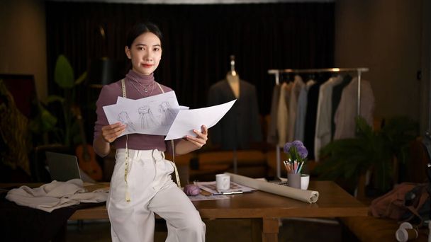 Egy profi és gyönyörű Y-generációs ázsiai női divattervező vagy szabó, aki az asztalra támaszkodik modellvázlataival.. - Fotó, kép