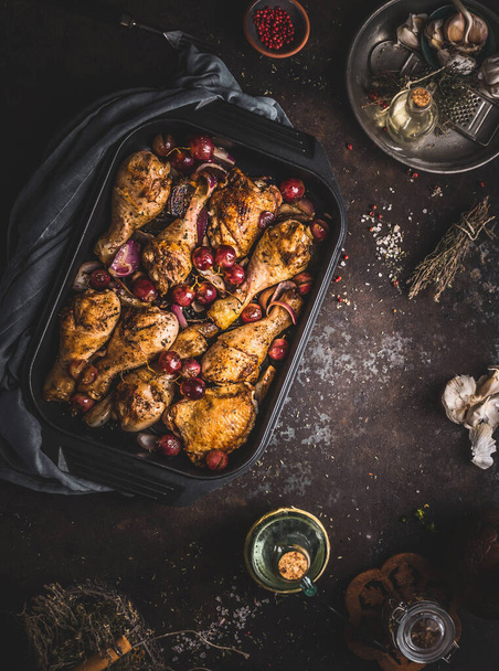 Tasty roasted chicken drumsticks with red onion and grapes in cast iron casserole pan on dark rustic background, top view - Fotografie, Obrázek