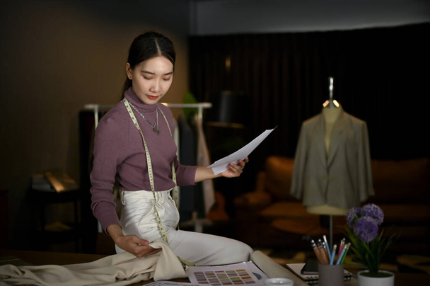 Attractive young Asian female tailor or fashion designer checking the quality of a new fabric and focuses on working on her new design in her studio. - Фото, зображення