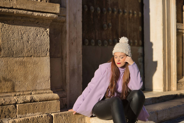 Young woman, beautiful, straight brown hair, leather pants, sweater, coat and wool hat, sitting on a step pensive and relaxed. Concept beauty, fashion, autumn, winter, cold. - Φωτογραφία, εικόνα