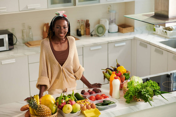 Ευτυχισμένο μαύρο γυναίκα στέκεται στο πάγκο της κουζίνας με φρέσκα παντοπωλεία έτοιμο να μαγειρέψουν υγιεινό γεύμα - Φωτογραφία, εικόνα