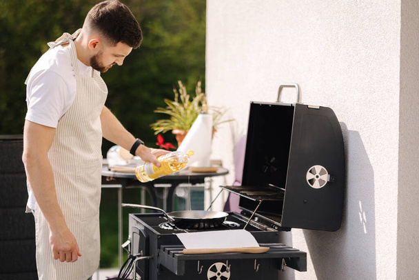 Handsome man add oil on frying pan. Preparing food outdoor. BBQ concept on terrace. High quality photo - Photo, Image